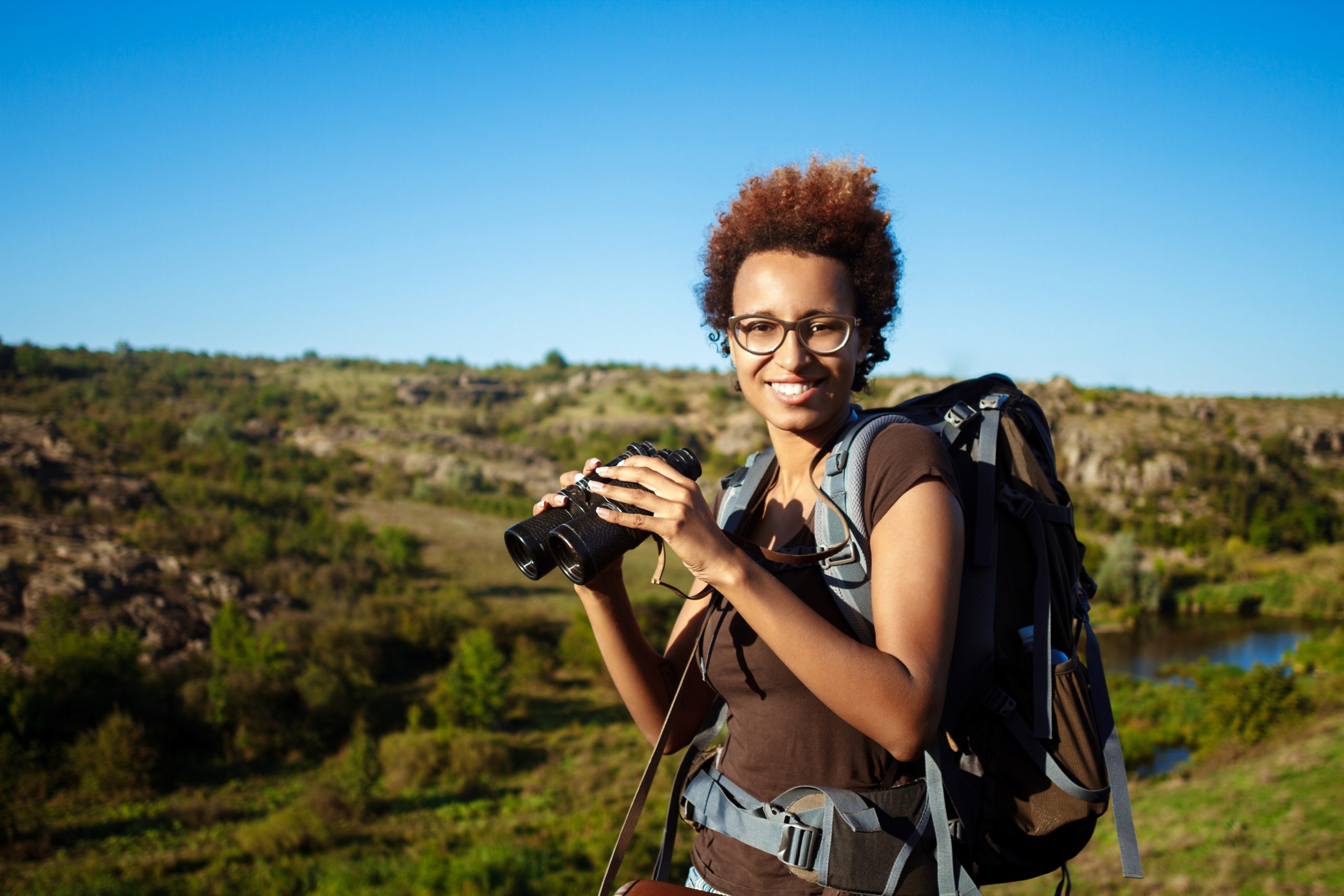 Young beautiful african girl with backpack smiling, looking at camera, holding binoculars, canyon background. Copy space.